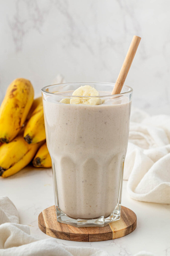 side view of banana boat smoothie in clear glass with straw and slices of banana on top on wooden coaster with bananas in background