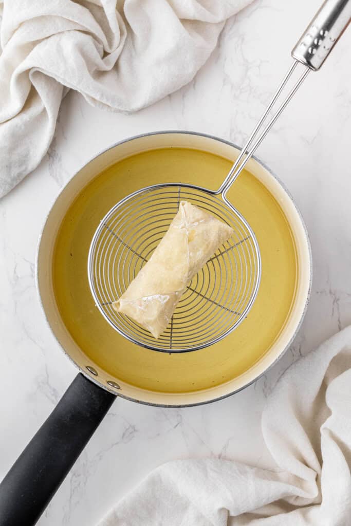 overhead view of egg roll getting dipped into hot oil with wire spoon