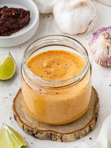 glass jar with sauce on wooden coaster with limes, peppers and garlic in the background