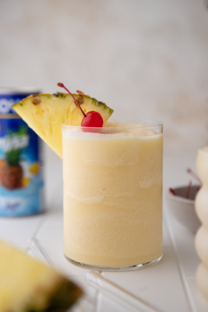 side view of topical Goya Pina colada with pineapple wedge and cherry with coconut cream in the background
