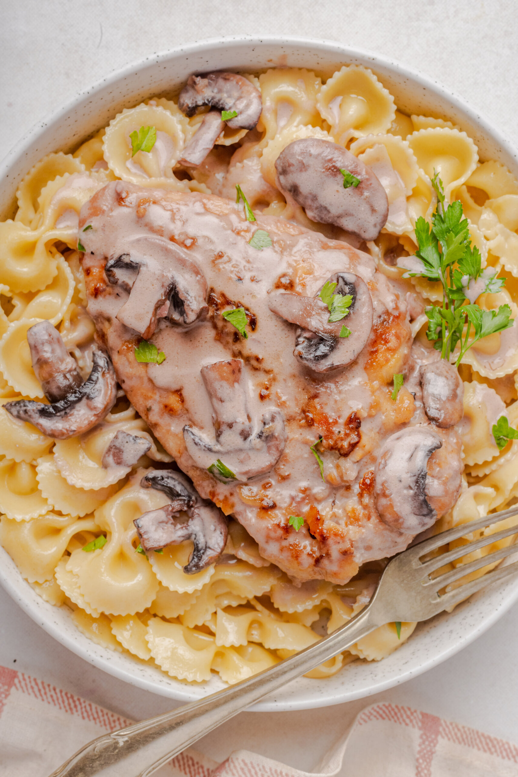 close up overhead view of copycat Cheesecake Factory Chicken Marsala on pasta with parsley and mushrooms with a white bowl with fork