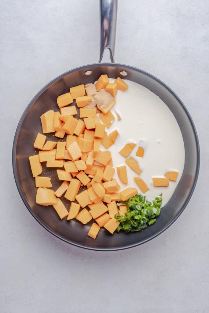 Chunks of cheese, cream, spices, and chopped peppers in a skillet before melting.