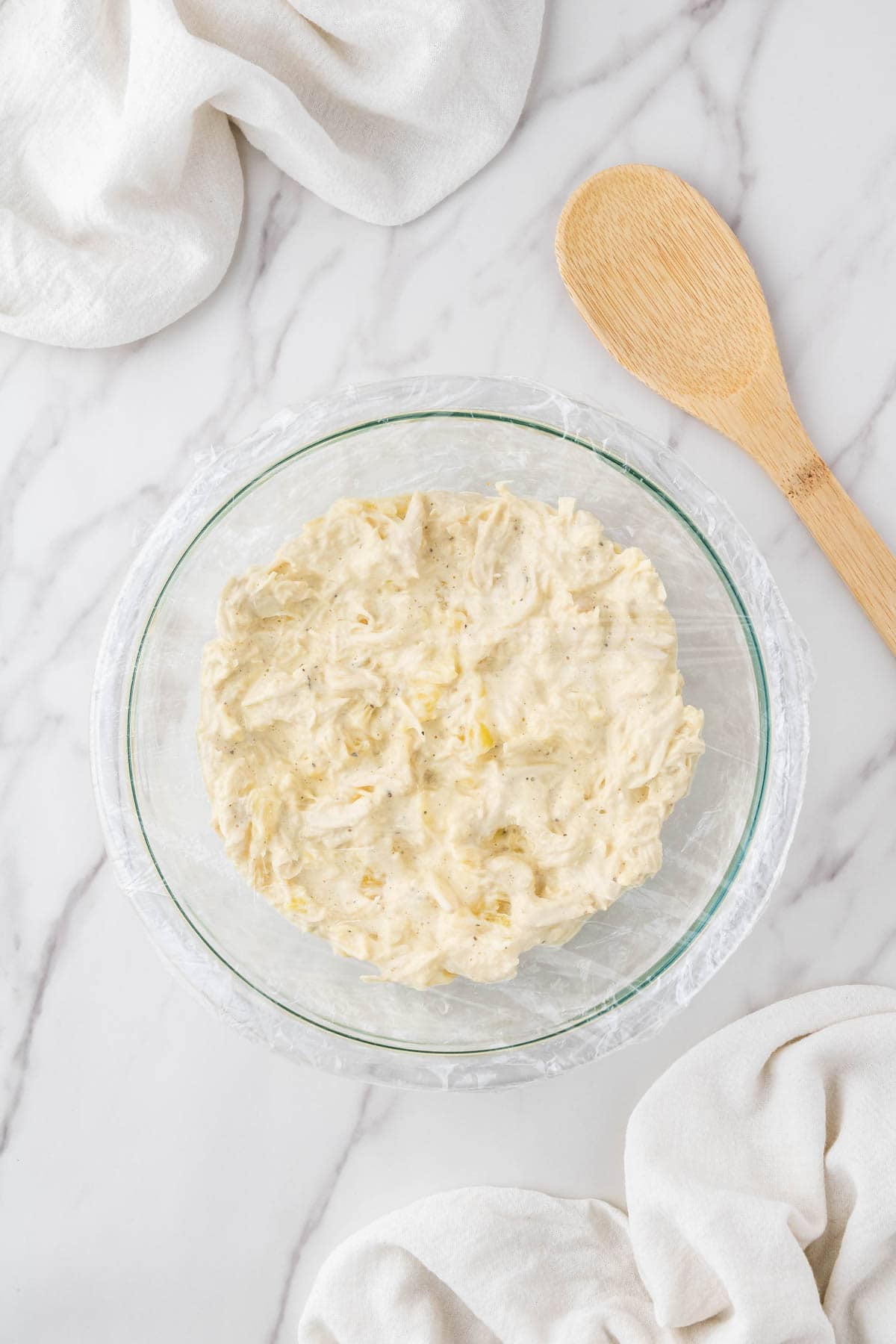 mixed ingredients in clear bowl with plastic wrap over the top of the bowl