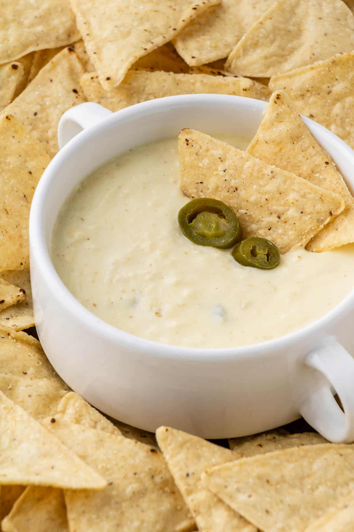 Moe's Queso recipe in white cup with jalapeños with a cup dipped in on a bed of chips