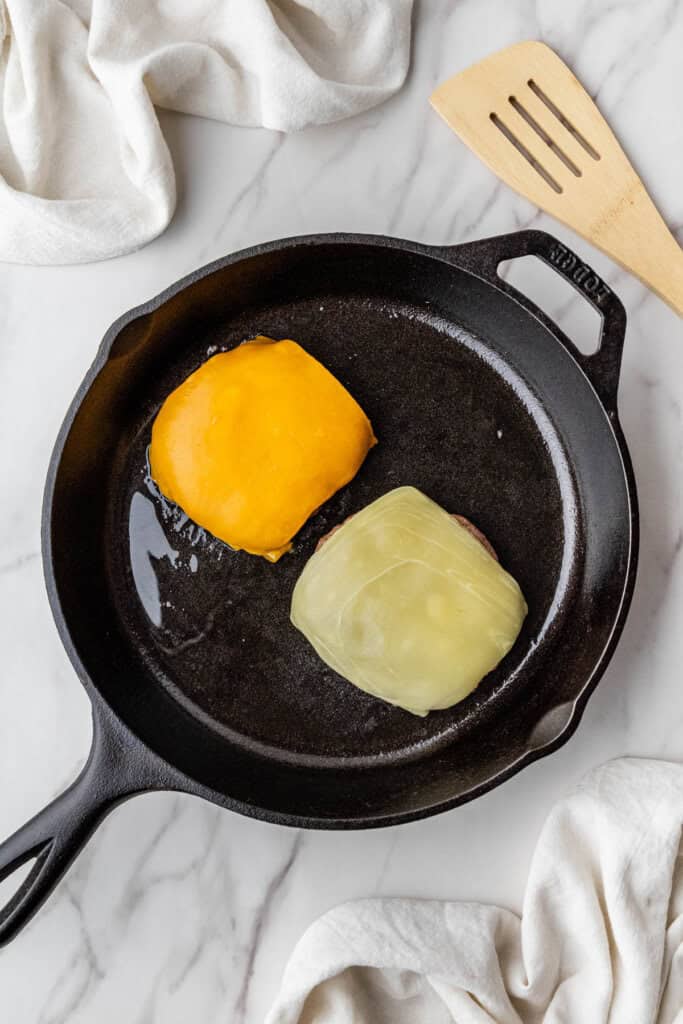 two hamburger patties with Swiss cheese and cheddar cheese on top in cast iron skillet
