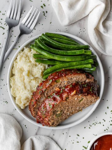 slices of Cracker Barrel Meatloaf recipe on a white plate with mashed potatoes and green beans