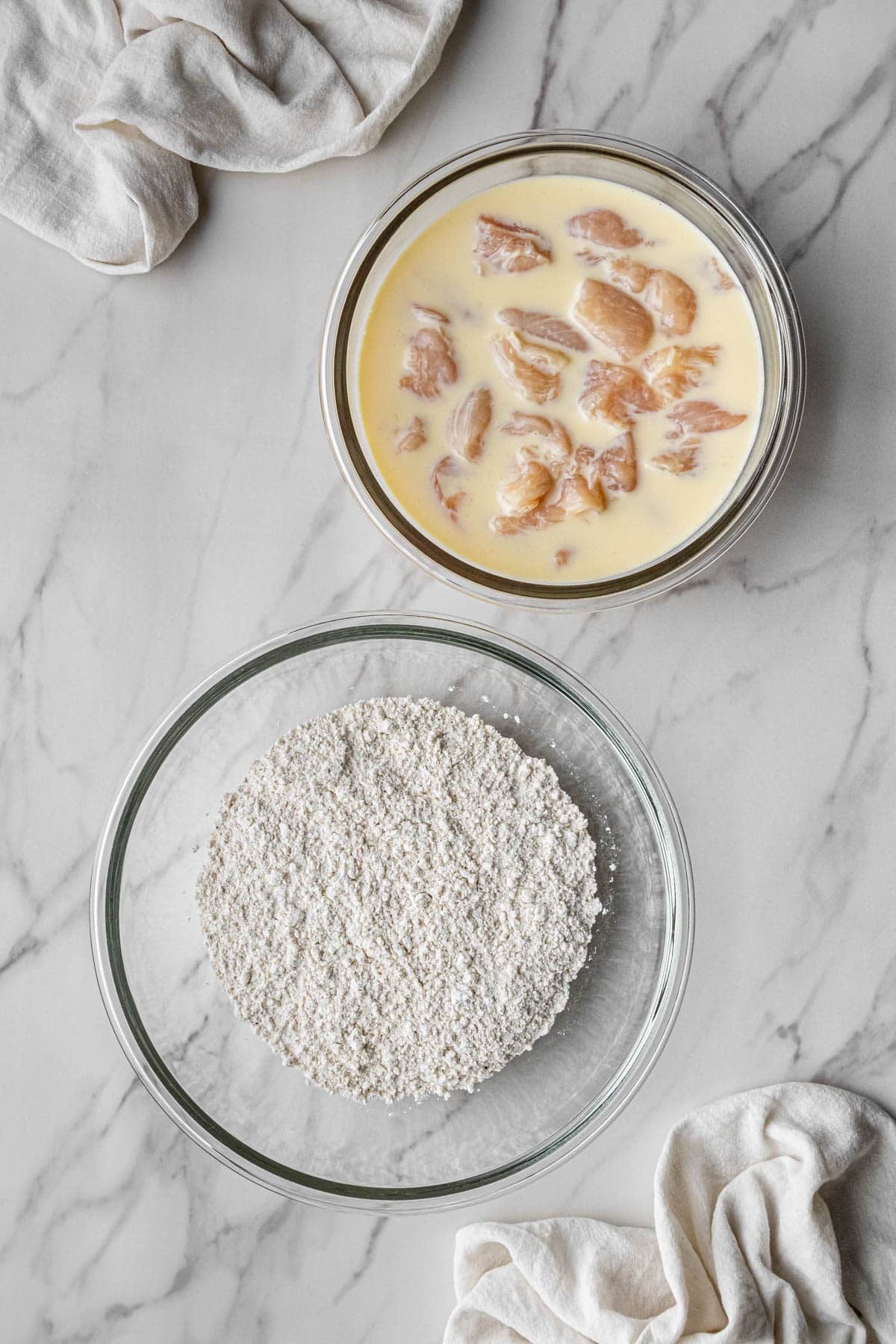 meat pieces in milk mixture in clear bowl with coating mixture in another bowl beside it