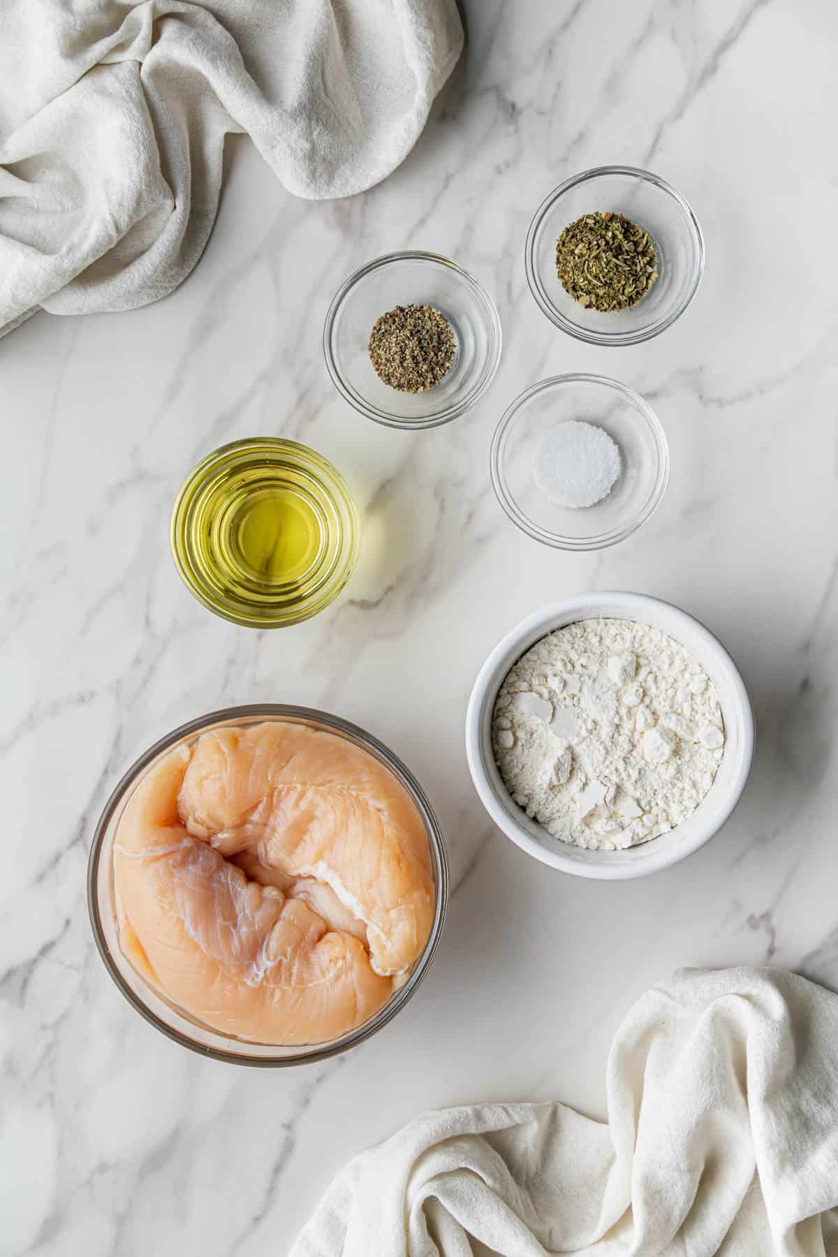 Flat lay shot of the ingredients for the pan-seared chicken breast for the olive garden Chicken Scampi recipe. There's chicken breasts, flour, oil, salt and pepper and herbs, all in separate glass bowls.