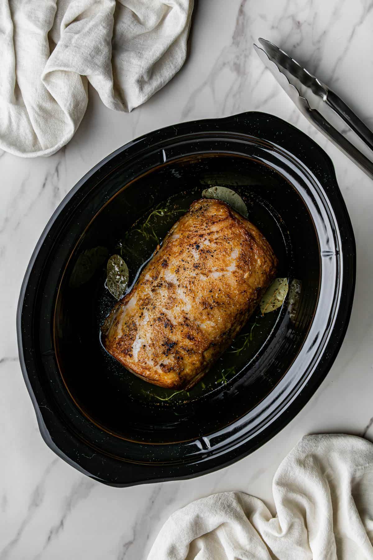 Flat lay shot of an open Crockpot, with the seared meat in it. Other ingredients such as chicken broth and bay leaves are poured into it as well.