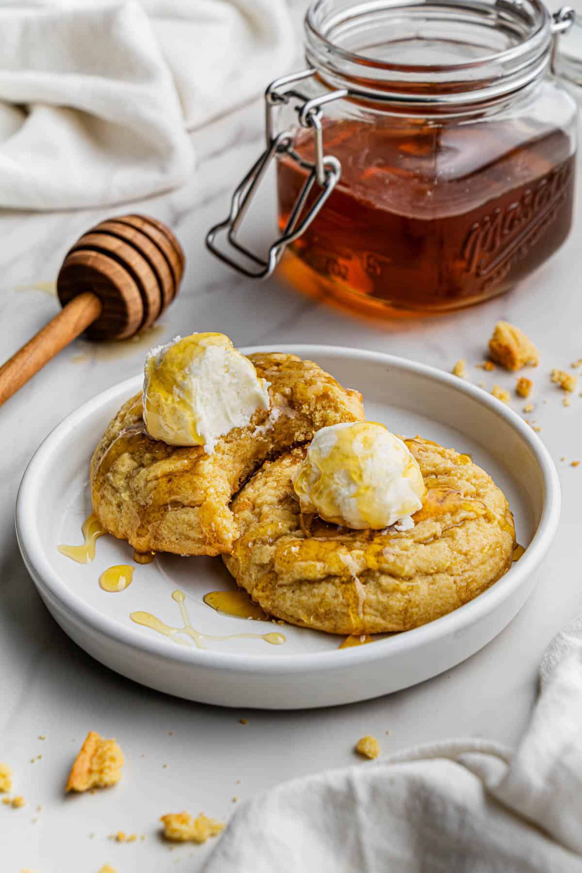 Two of the finished Crumbl Cornbread Cookies Copycat recipe on a white ceramic plate, both drizzled with honey, and with one of the cookies (on the left) having been bitten into.