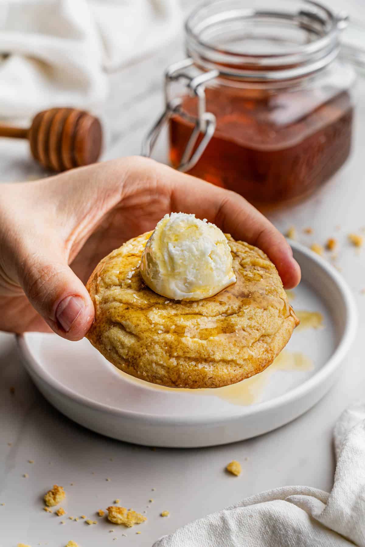 A hand holding a Crumbl Copycat Cornbread Cookie with a ceramic plate behind it.