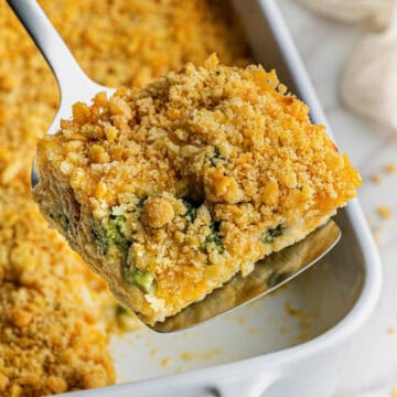 A square slice of Copycat Cracker Barrel Broccoli Cheese casserole recipe being lifted from a white baking dish with a spatula.