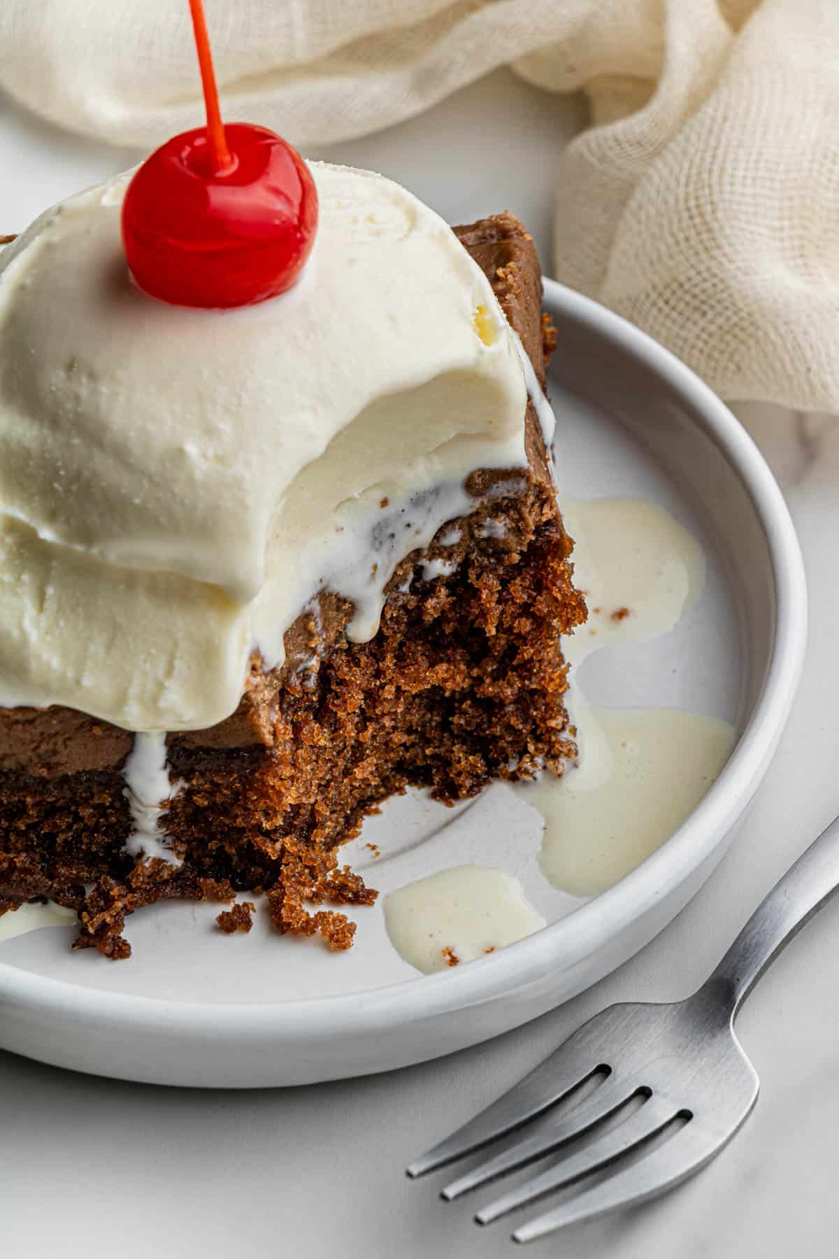 High angle shot of a slice of the Cracker Barrell Coca cola Cake recipe with a scoop if vanilla ice cream with a cherry on top. I piece of cake has been taken out of the tip. Beside the ceramic plate where the cake is, is a fork.