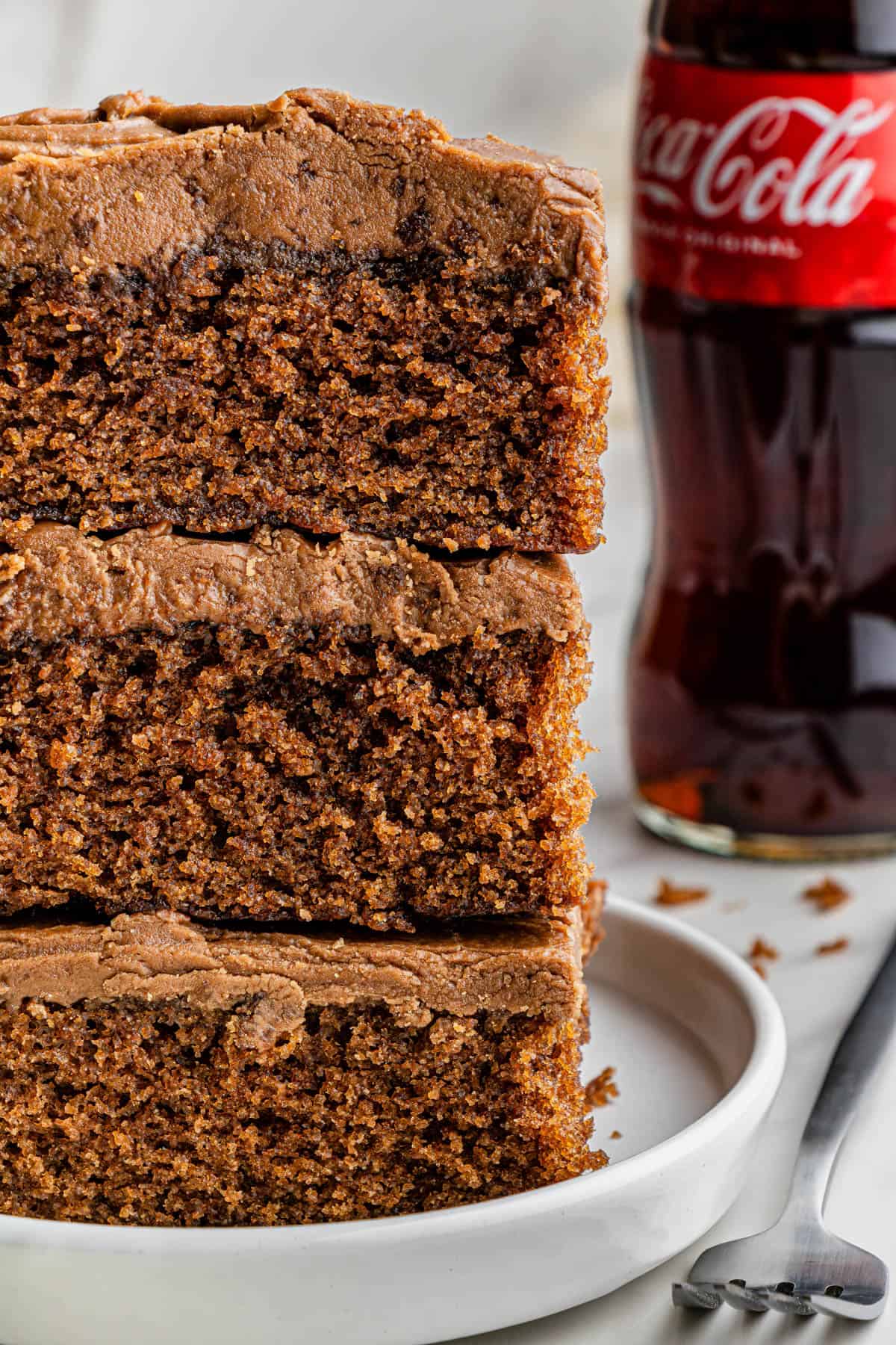 Partial shot of 3 slices of the finished Cracker Barrel Copycat Coca-Cola Cake stacked on a white ceramic plate. Behind the cake stack is a bottle of regular Coke.