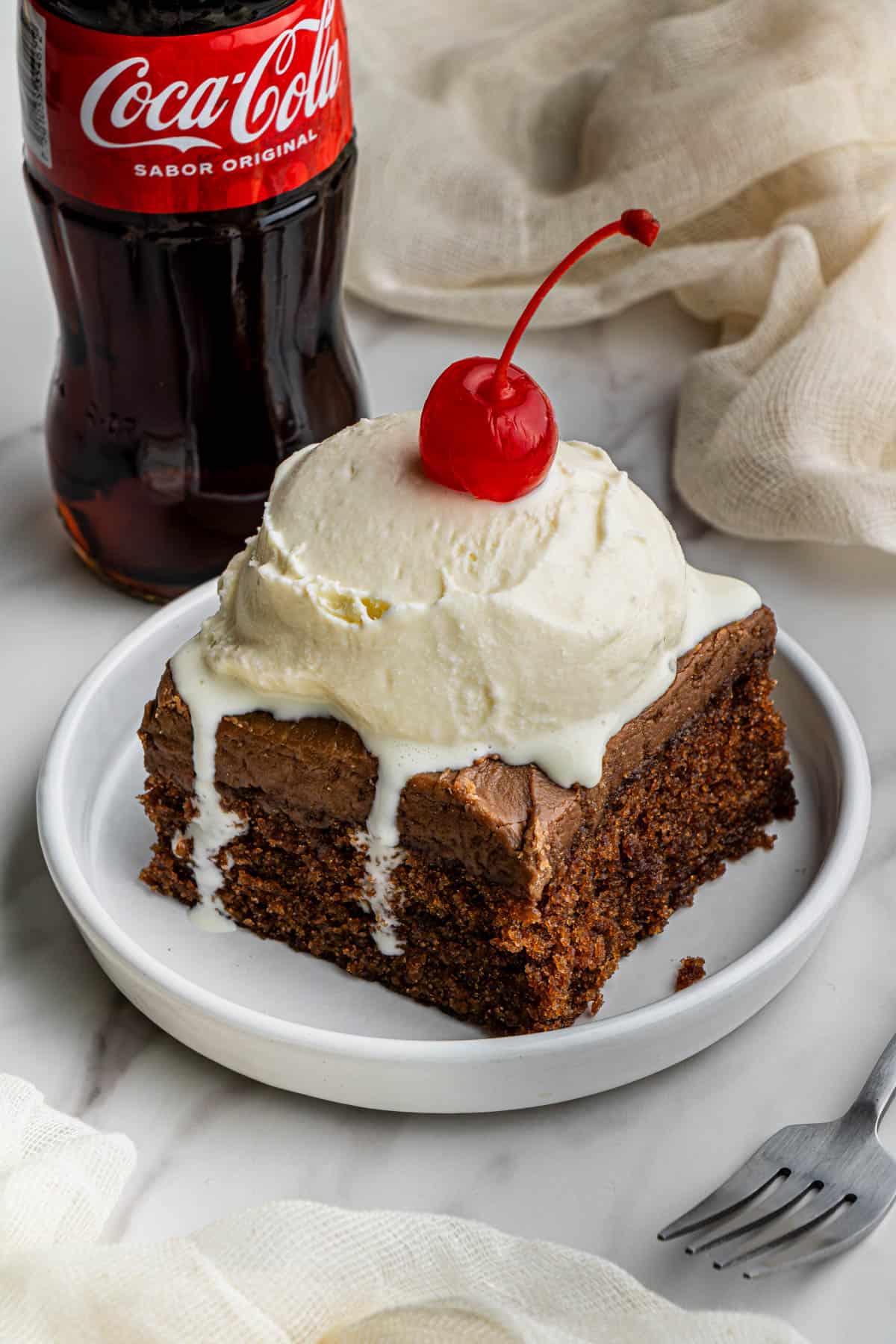 A slice of the fudgy cracker barrel Coca-Cola Cake recipe topped with a slice of vanilla cake with a cherry on top. The dessert is on a white ceramic plate with a bottle of regular Coke beside it.