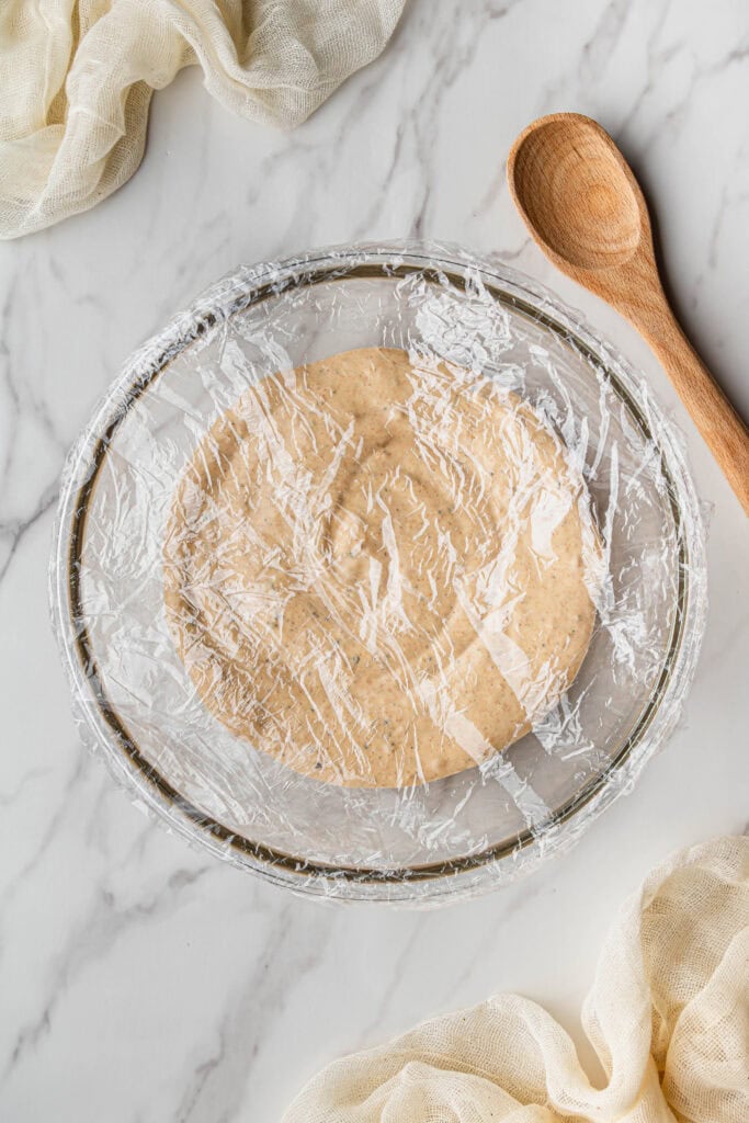 Flat lay shot of a glass bowl with the mixed ingredients, covered with plastic wrap and placed on a marble surface.