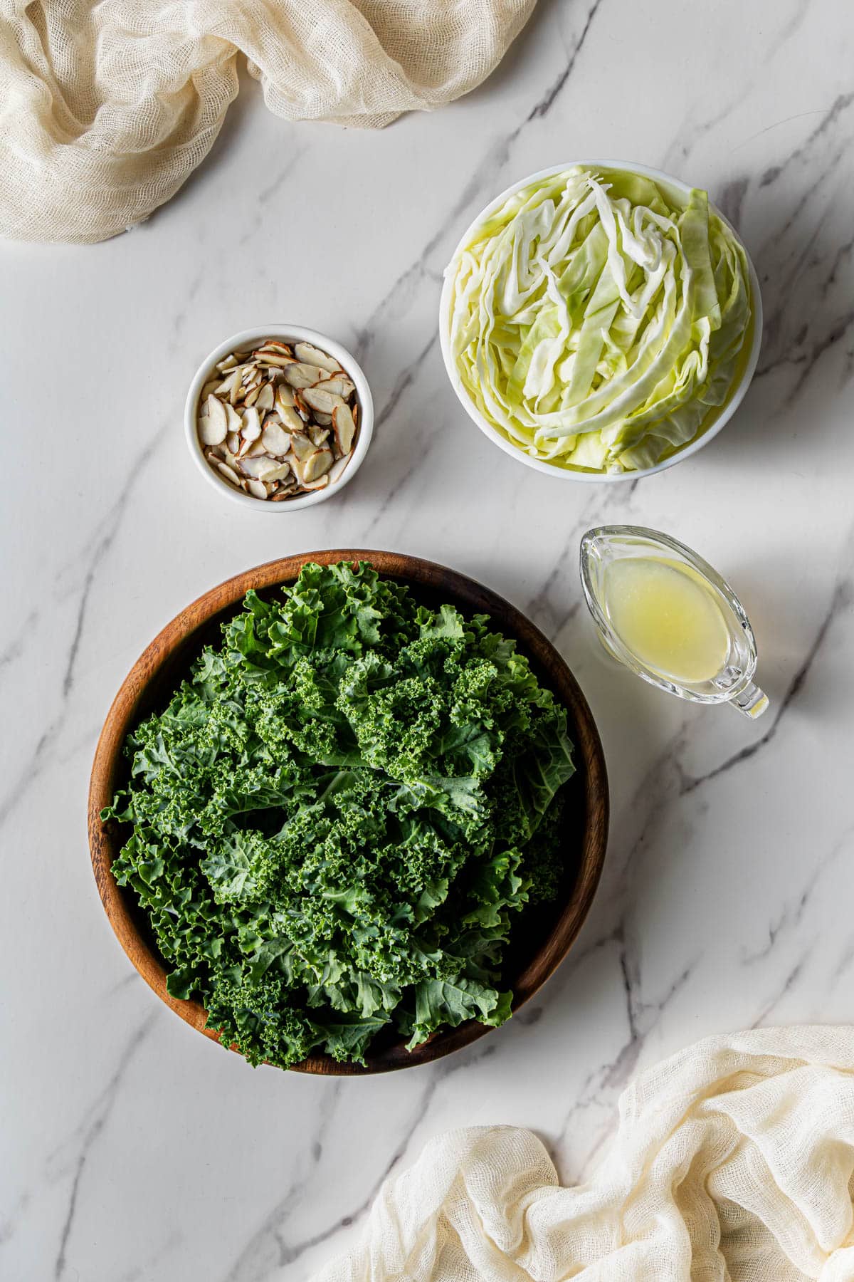 Flat lay shot of ingredients such as curly kale in a wooden bowl, dressing in a small gravy boat, sliced almonds in a bowl, and cabbage in another bowl.