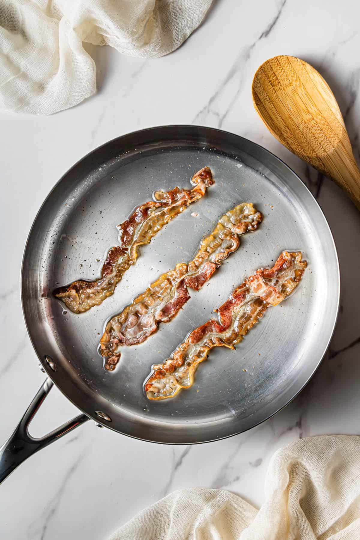 Flat lay shot of 3 slices of bacon rendering their fats in a skillet.
