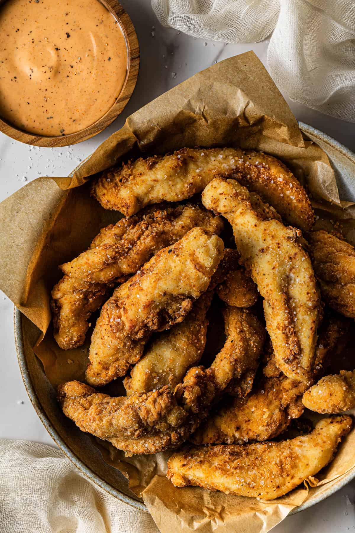 Close-up of crispy homemade chicken tenders in a parchment-lined bowl with coarse salt on top and a dipping sauce on the side.