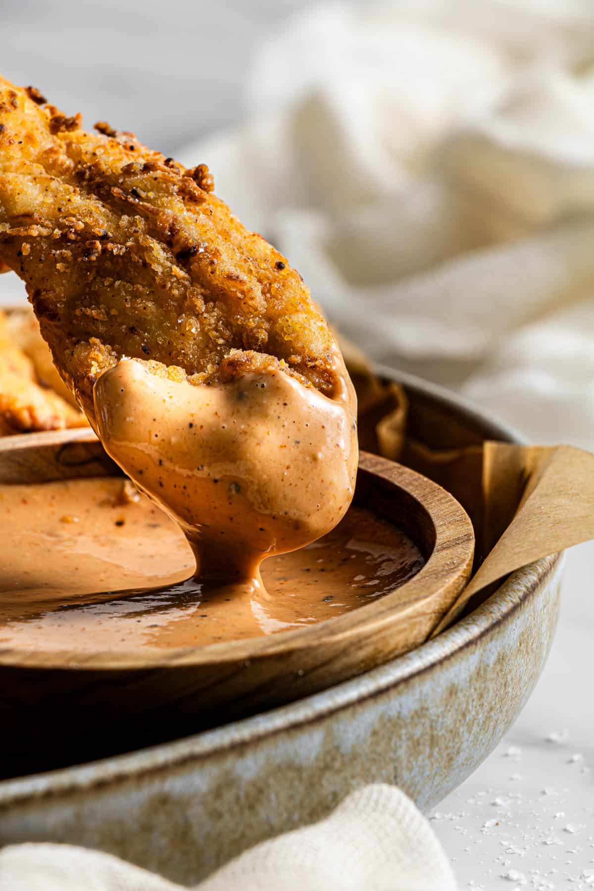 Close-up of a crispy fried chicken tender being dipped into a creamy seasoned sauce.