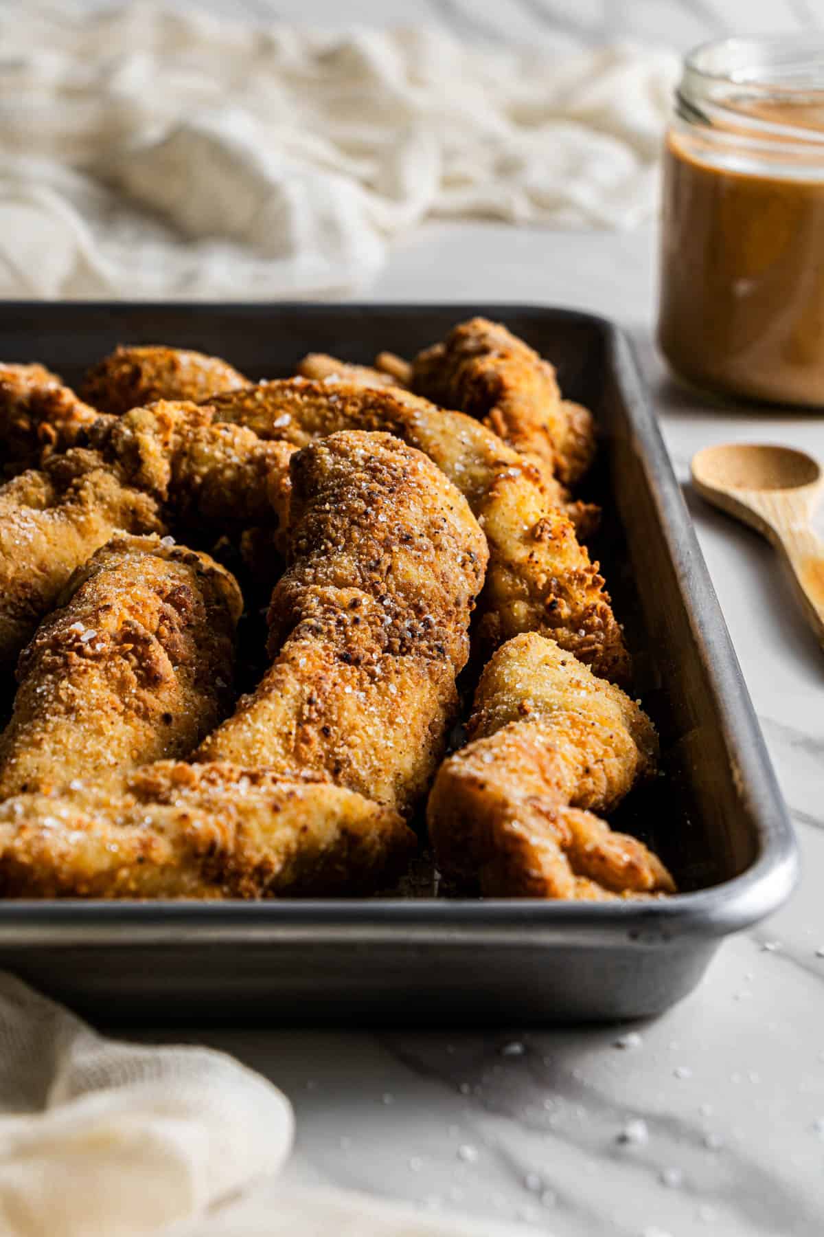 Close-up view of crispy chicken tenders in a baking tray with a jar of dipping sauce in the background.