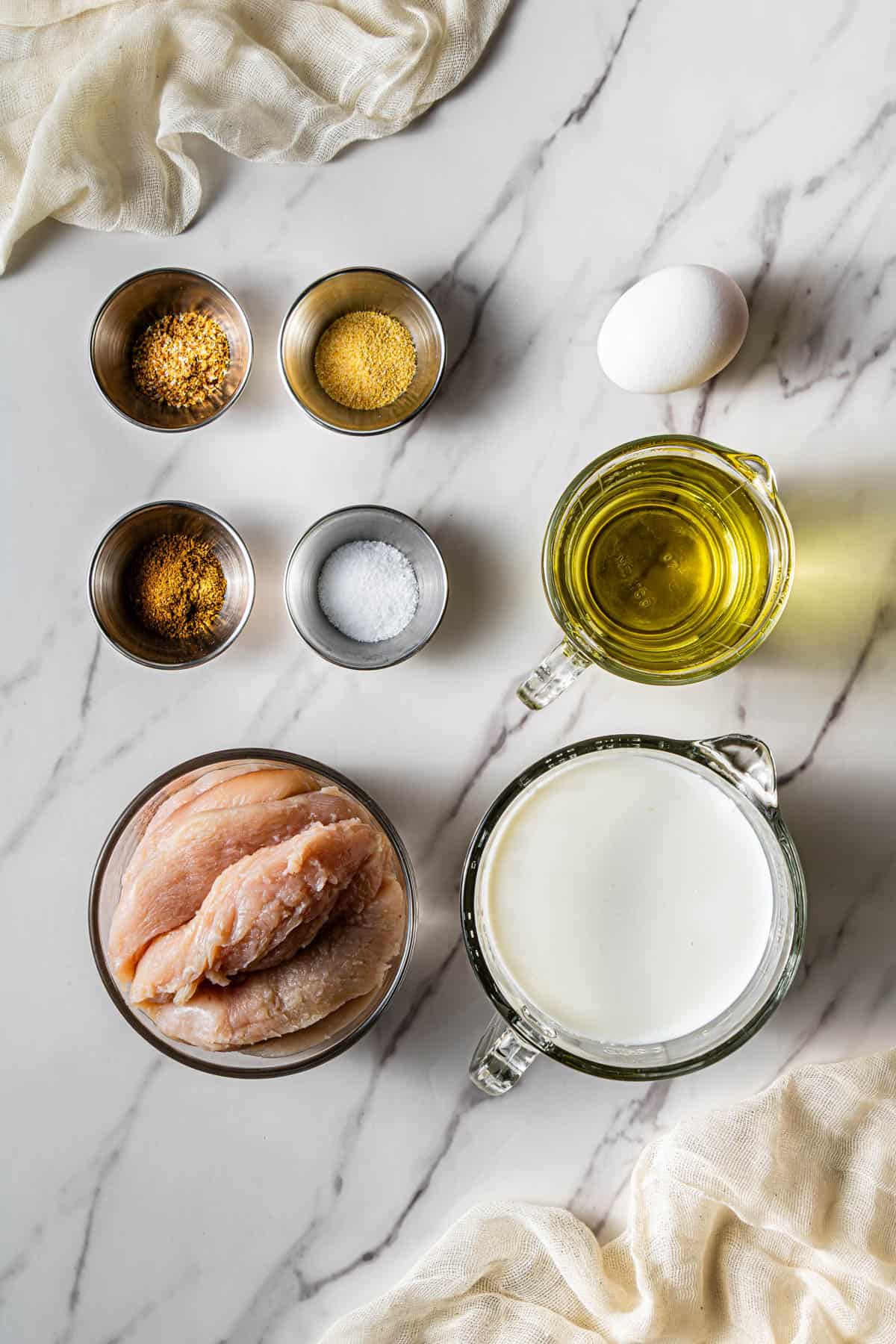 Overhead view of raw chicken tenders, eggs, buttermilk, oil, and seasoning ingredients arranged on a marble surface for making homemade chicken tenders.