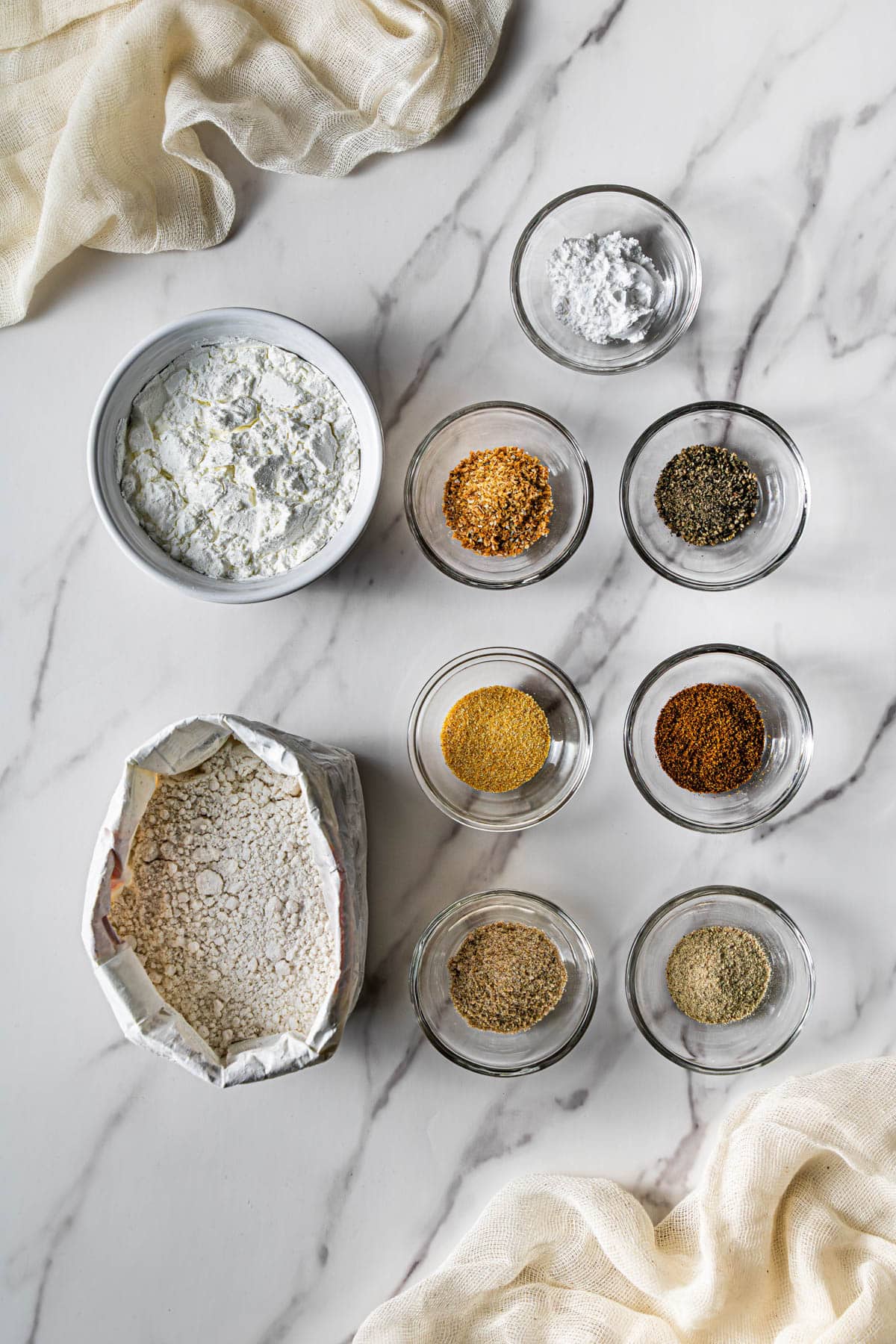 Flat lay of seasoned flour mixture with bowls of spices, pepper, garlic seasoning, and baking powder on a marble surface for coating chicken tenders.