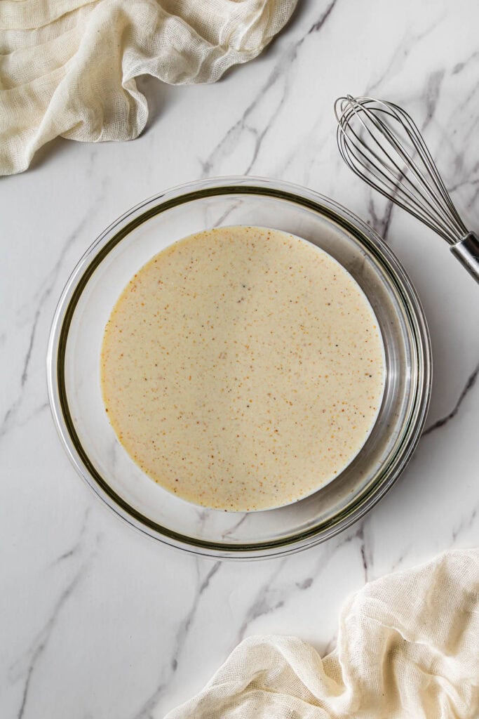 Glass bowl filled with seasoned buttermilk marinade on a marble surface with a whisk beside it.