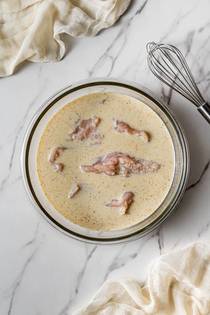 Raw chicken tenders soaking in a seasoned buttermilk marinade inside a glass bowl.