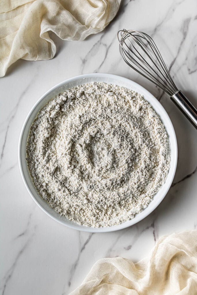 Bowl filled with a seasoned flour mixture on a marble surface with a whisk beside it.