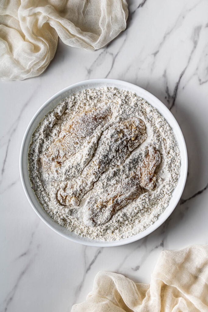 Raw chicken tenders being coated in a seasoned flour mixture in a shallow bowl.