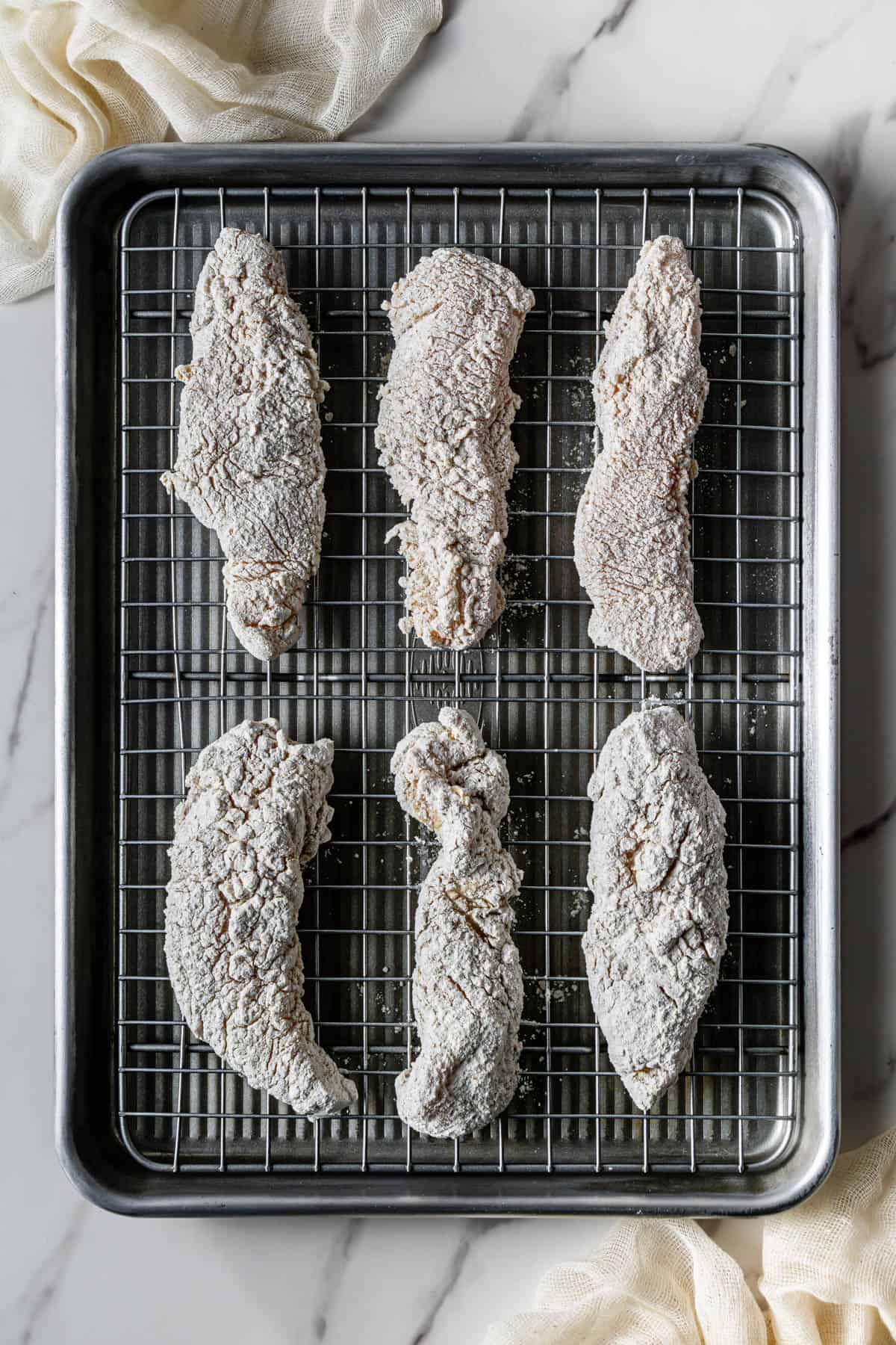 Raw chicken tenders coated in flour resting on a wire rack before frying.