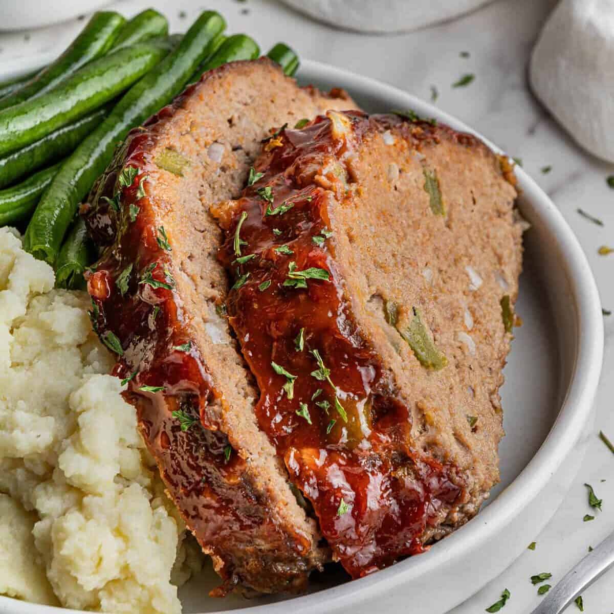 slices of Cracker Barrel Meatloaf recipe on a white plate with mashed potatoes and green beans