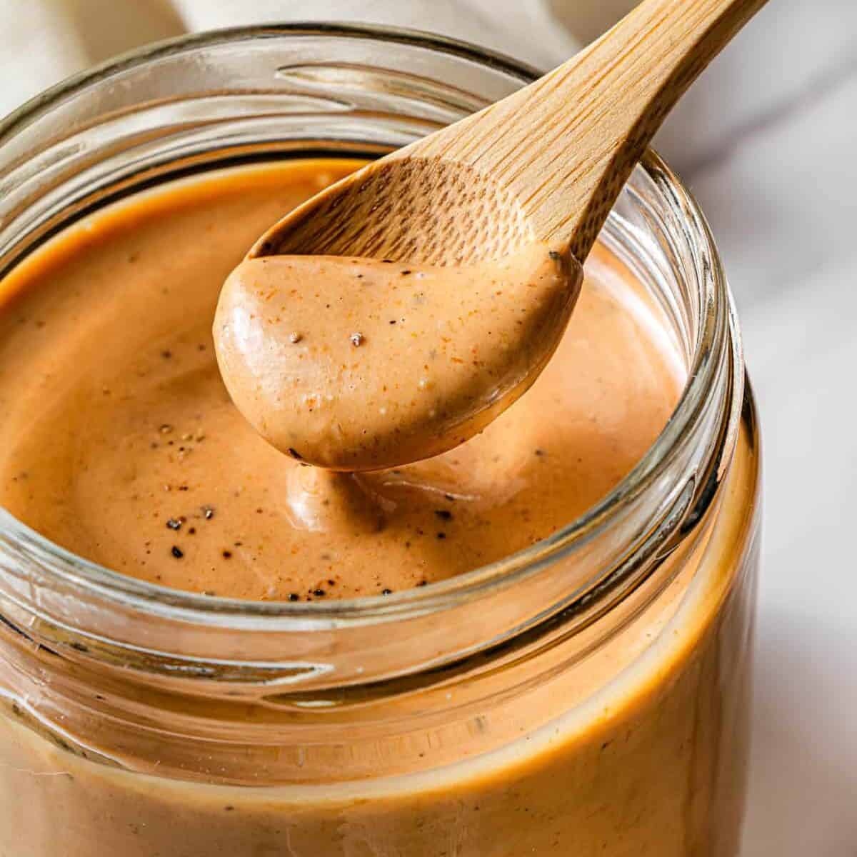 A glass jar with the Raising Cane sauce in it, and a small wooden spoon scooping up a bit of the sauce.