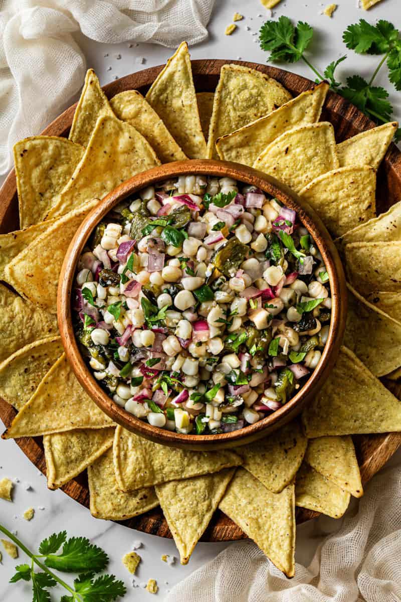 Chipotle corn salsa recipe in a wooden bowl surrounded by tortilla chips on a round board
