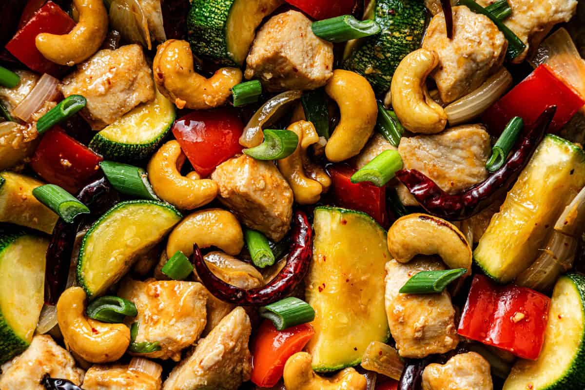 Close-up of kung pao chicken with chicken thighs, cashews, zucchini, red bell pepper, scallions, and dried chilies coated in kung pao sauce