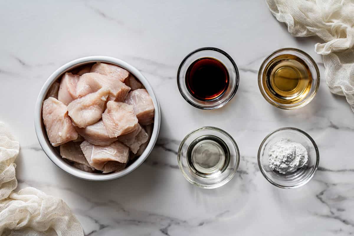 Chicken thigh pieces in a white bowl with soy sauce, Shaoxing wine, oil, and cornstarch in small glass bowls