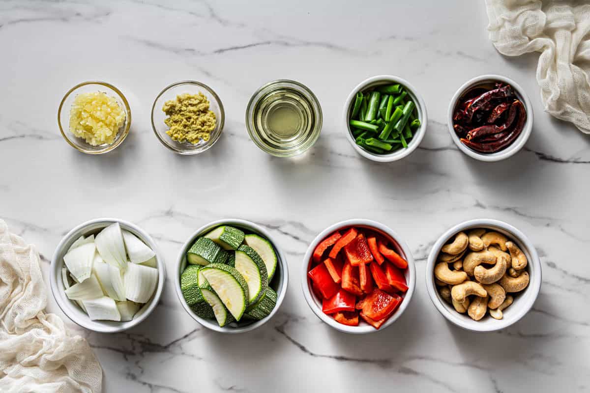 Prepped kung pao chicken vegetables including zucchini, red bell pepper, onion, cashews, scallions, dried chilies, garlic, and ginger in small white bowls