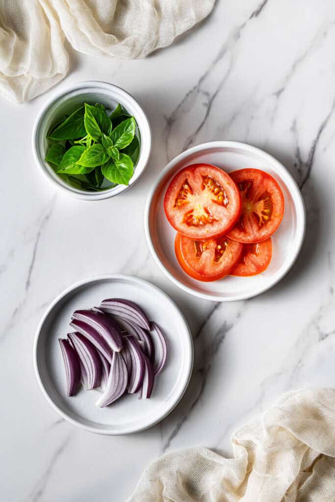 Sliced tomatoes, basil leaves, and red onion prepared for chicken panini filling